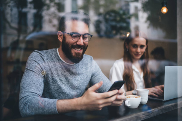 Bearded man in casual clothes and eyeglasses sitting at cafe table smiling and browsing smartphone next to girlfriend typing on laptop and looking at phone screen - Valokuva, kuva
