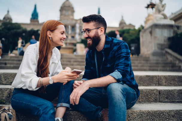Happy youthful couple looking at each other and smiling while sitting on stairs in front of university and resting after studies - Zdjęcie, obraz
