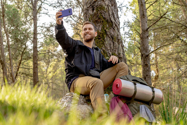 Jongeman wandelen de berg op met een selfie in het bos glimlachend, man in kampeerkleding en kampeerrugzak - Foto, afbeelding