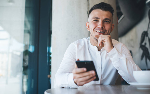 Positive smiling male wearing formal clothes and browsing smartphone while sitting at table with cup of coffee in modern cafe and looking at camera - Фото, зображення