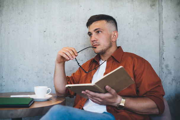 Thoughtful male freelancer in casual outfit thinking about notes in daily planner while drinking coffee in street cafeteria and looking away - Photo, Image