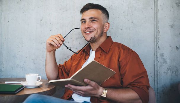 Молодой человек в повседневной одежде улыбается в камеру и снимает очки после чтения книги во время отдыха в уютном кафе - Фото, изображение