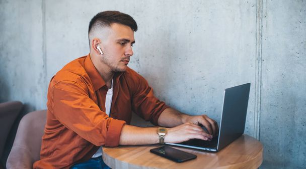 Side view of young male in casual wear with earphones sitting at table with smartphone and using laptop while messaging on social media - Фото, зображення