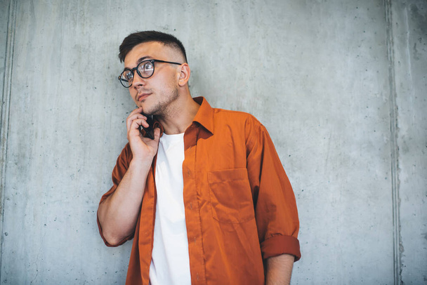 Serious young male in casual clothes and eyeglasses standing against gray wall and looking away while waiting answer to phone call - Fotografie, Obrázek