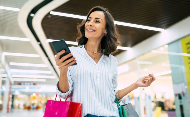 Modern gorgeous young smiling woman in trendy stylish clothes with bright colorful shopping bags is using her smart phone while walking in the mall - Photo, Image