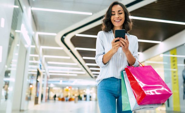 Modern gorgeous young smiling woman in trendy stylish clothes with bright colorful shopping bags is using her smart phone while walking in the mall - Foto, Imagem