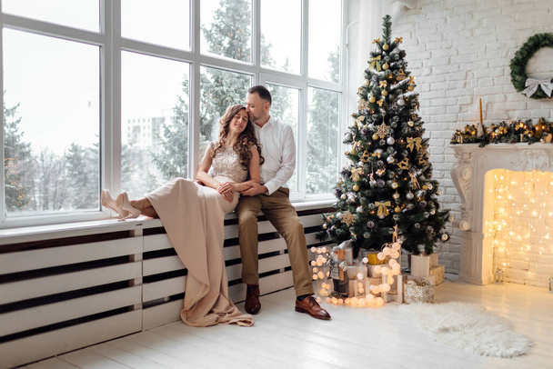 ζευγάρι ερωτευμένο σε γιορτινά ρούχα κάθεται και αγκαλιάζει κοντά στο μεγάλο παράθυρο και χριστουγεννιάτικο δέντρο σε διακοσμημένο στούντιο. Καλές διακοπές. - Φωτογραφία, εικόνα