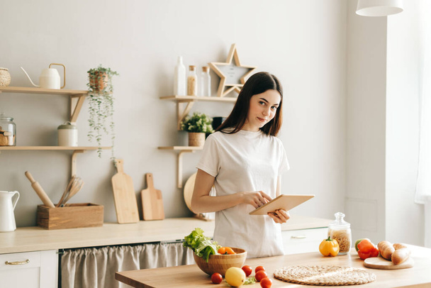 Young caucasian woman use tablet computer in the modern kitchen, preparing salad, read recipe. - Foto, Imagem