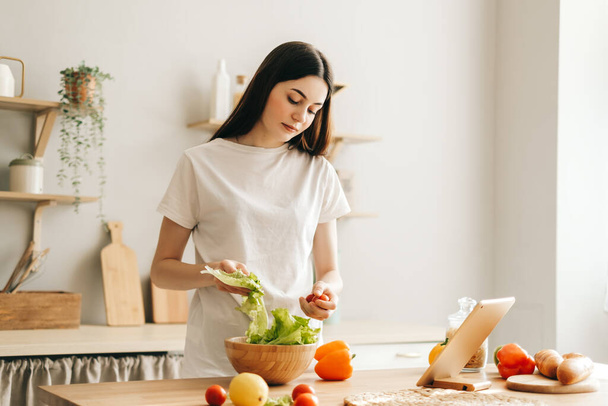 Young caucasian woman use tablet computer in the modern kitchen, preparing salad, read recipe. - Foto, Imagem