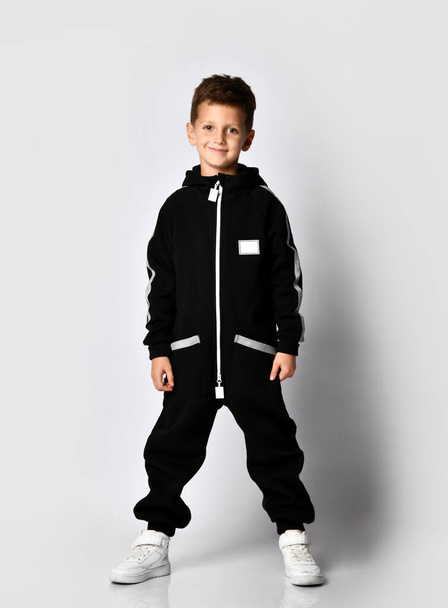 Little brunette kid in colorful tracksuit and sneakers on a gray background. - Foto, Bild