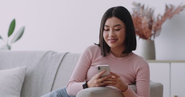 Nadšený asijské dáma chatování na mobilu, čtení radostné zprávy, odpočívá na pohovce doma - Záběry, video