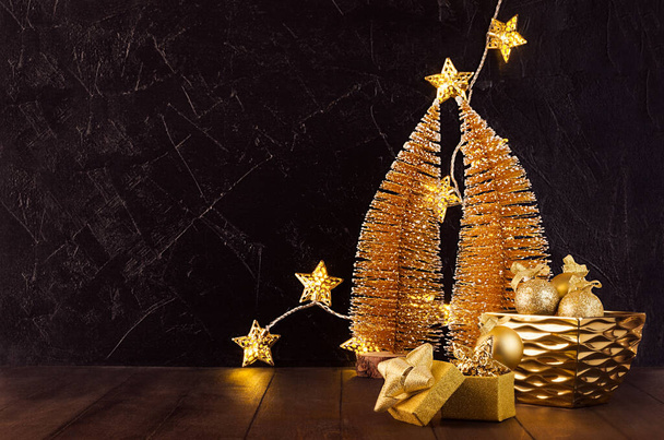 New Year home decor in dark and golden color - christmas trees, glowing lights, stars, balls, gift box, ribbons in black interior on wood desk, copy space. - Foto, imagen