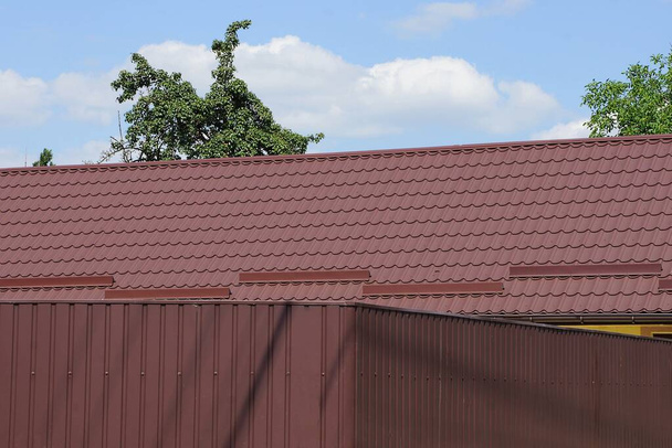 красная черепичная крыша дома за коричневым металлическим забором стены против голубого неба - Фото, изображение