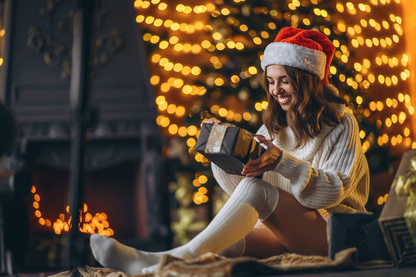 Christmas, New Year. Pretty woman in warm sweater, socks and christmas hat, sitting on the floor at home with gift boxes and new year tree and lights behind - Foto, immagini