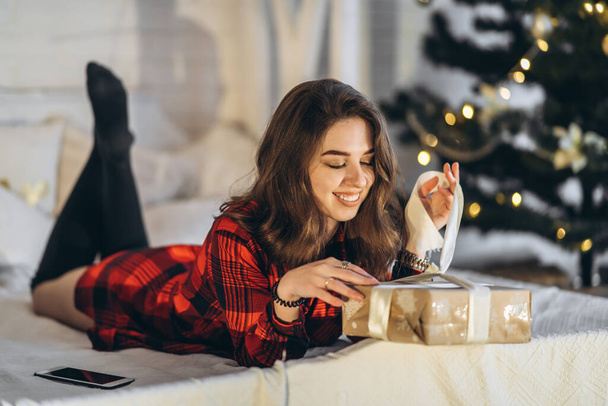 Christmas, New Year. Pretty woman in shirt and socks lying in bed with christmas gift box, new year tree behin - Foto, imagen