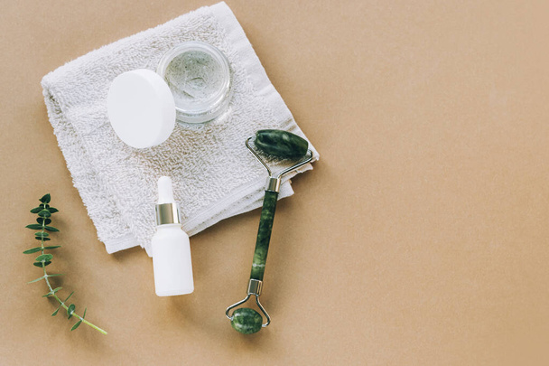 Kosmetikserum in weißer Markenflasche, Sahneglas und Gesichtswalze. Handtuch, Eukalyptusblätter auf beigem Hintergrund. Draufsicht, flache Lage, Kopierraum. - Foto, Bild