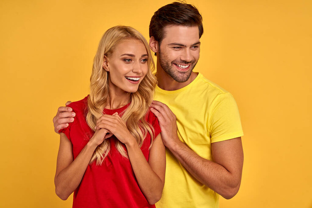 Улыбающаяся молодая пара друзей девушка в красно-желтых футболках позирует изолированно на фоне желтой стены студийный портрет. Люди эмоции образ жизни концепции. Нарисуй копировальное пространство. Обнимаясь, оглядываясь в сторону - Фото, изображение