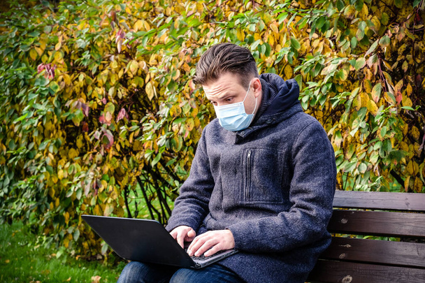 Bello giovane europeo in un parco con un computer portatile con una maschera medica. Freelance che lavora fuori dall'ufficio durante un'epidemia di Covid-19. Focus selettivo - Foto, immagini