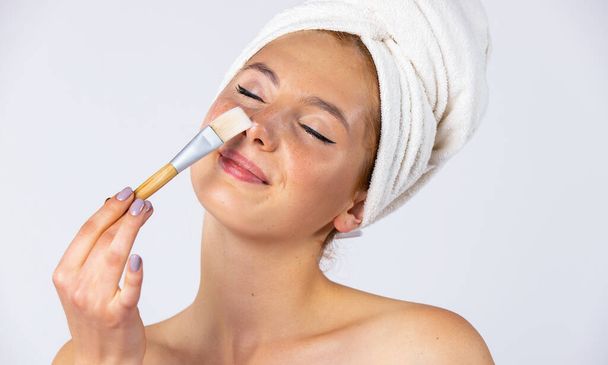 A girl with a towel on her head and hangs on her face, applies a gel mask with a brush. Fashionable female model with beautiful appearance. Photo on white background. High quality photo - Φωτογραφία, εικόνα
