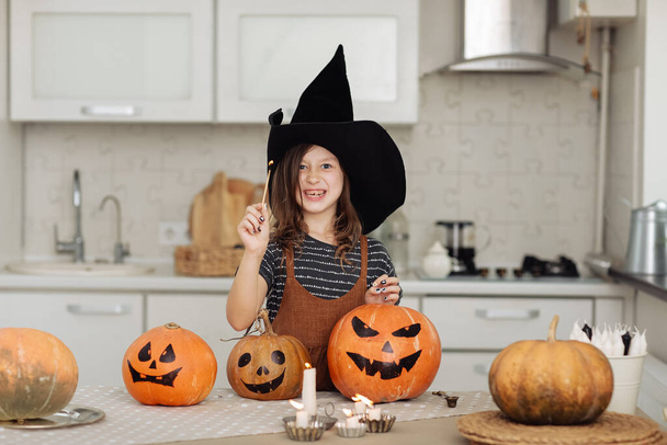 Boldog Halloweent! Aranyos kislány boszorkányjelmezben, faragott sütőtökkel. Boldog család készül Halloweenre. lány gyertyákat gyújt. - Fotó, kép