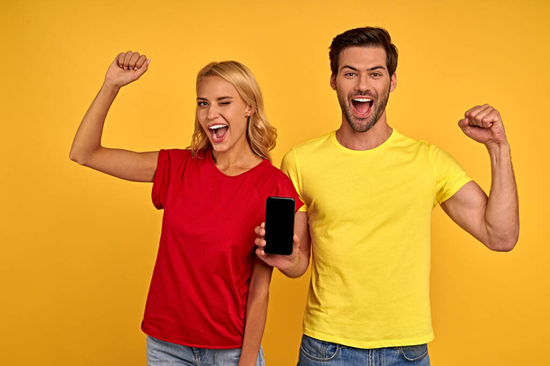 Joyful young couple two friends guy girl in casual clothes isolated on yellow background. People lifestyle concept. Mock up copy space. Hold mobile phone with blank empty screen, doing winner gesture. - Foto, immagini
