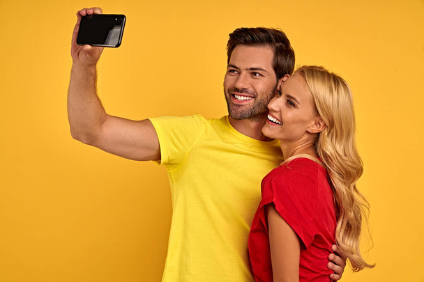 Cheerful couple two friends guy girl in red yellow t-shirts isolated on yellow background. People lifestyle concept. Mock up copy space. Doing selfie shot on mobile phone hugging - Photo, Image