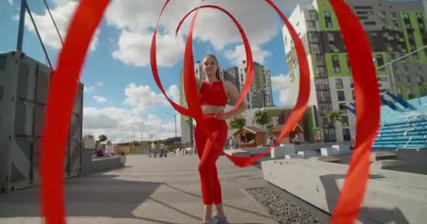 Молодая женщина в красном выполняет callisthenics с лентой на площадке тренировки на открытом воздухе, гимнаст делает акробатические упражнения в резиденциальной области, фитнес в городской среде, 4k 120p Прорес HQ - Кадры, видео