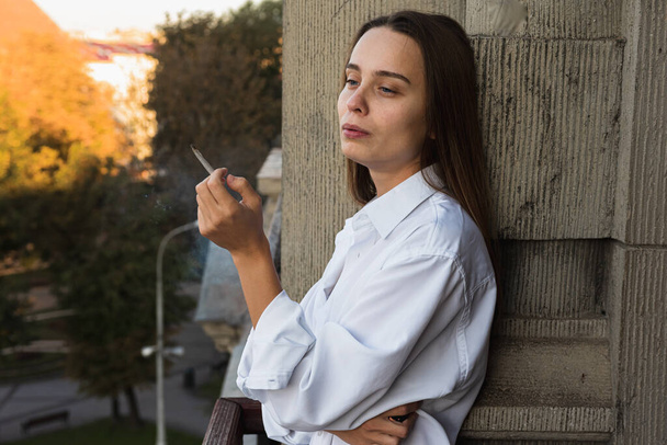 junges hübsches Mädchen raucht auf dem Balkon, Morgensonnenaufgang, kein gesunder Lebensstil - Foto, Bild