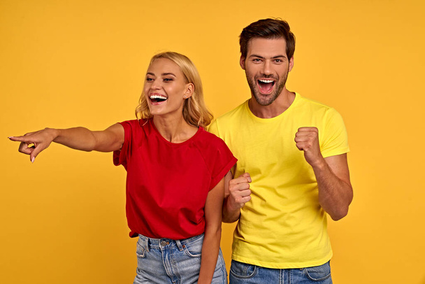 Glückliches junges Paar zwei Freunde Kerl und Mädchen in lässiger Kleidung posieren isoliert auf gelbem Hintergrund. Menschen Lifestyle-Konzept. Kopiertastatu.Zeigefinger zur Seite zeigen, Mund mit Händen bedecken. - Foto, Bild
