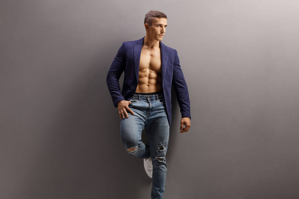 Male model in a suit shirtless with ripped jeans leaning on a gray wall  - Фото, изображение