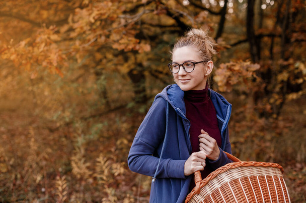 Chica con gafas en el bosque de otoño. Una mujer con una cesta camina por el bosque. Caminata de otoño. - Foto, imagen