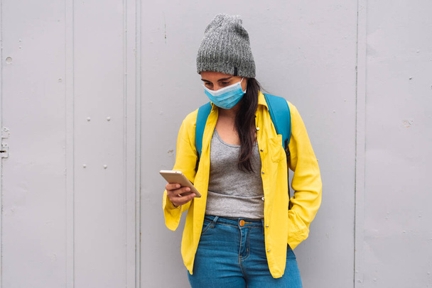 Stock photo of happy girl wearing yellow jacket using her smartphone in the street with face mask. - Zdjęcie, obraz