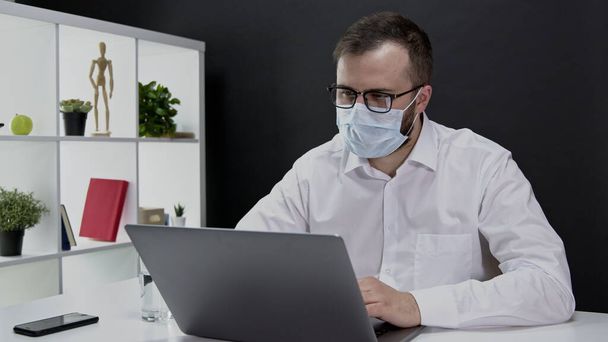 Красивый бизнесмен в защитной маске работает удаленно за ноутбуком на карантине - Фото, изображение