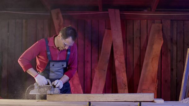 Αρσενικό ξυλουργός καθαρίζει την επιφάνεια της ξύλινης δέσμης χρησιμοποιώντας ηλεκτρικό πλάνη στο εργαστήριο - Φωτογραφία, εικόνα