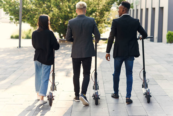 Professional multi ethnic co workers hold electric scooters walking near modern building and talking - Foto, imagen