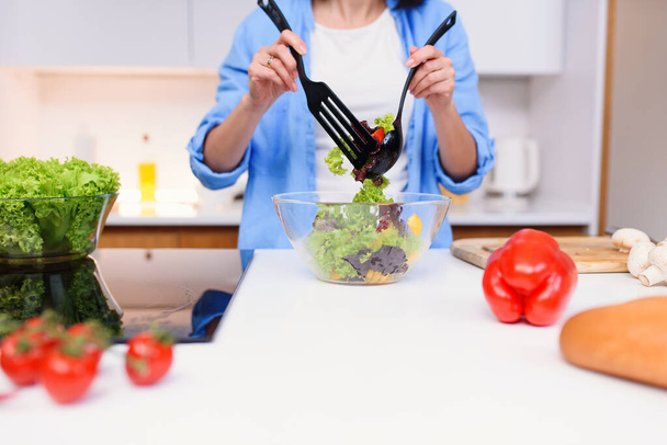 Close up photo of young womans hands mixing fresh vegetables vegetarian salad in a glass bowl at the kitchen. - 写真・画像