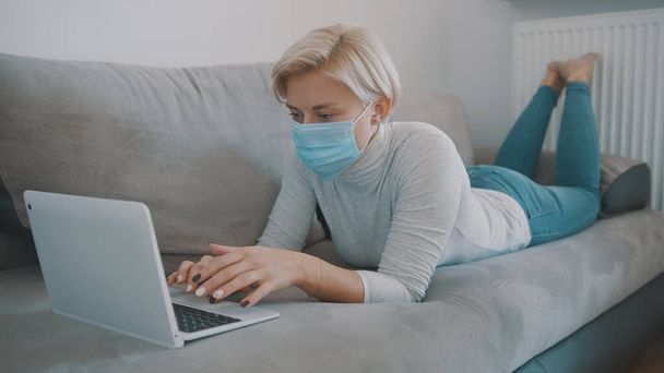 医療マスクをした若いブロンドの女性が遠隔操作で働いている。ソファに横になってノートパソコンを使う - 写真・画像
