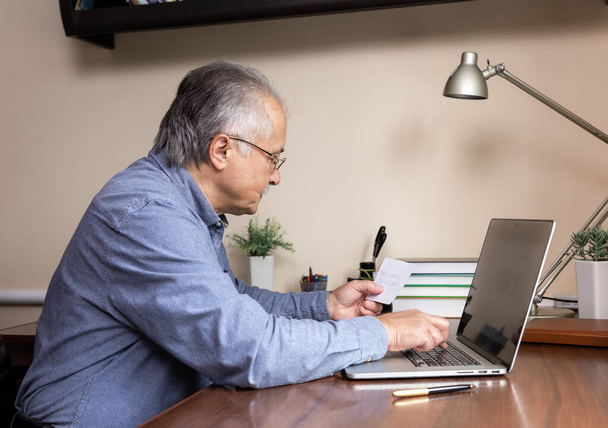 Старший научится пользоваться компьютером. Старик в стеклянной и синей рубашке вводит пароль с листа бумаги. Концепция использования ноутбука для онлайн-обучения в домашнем офисе - Фото, изображение