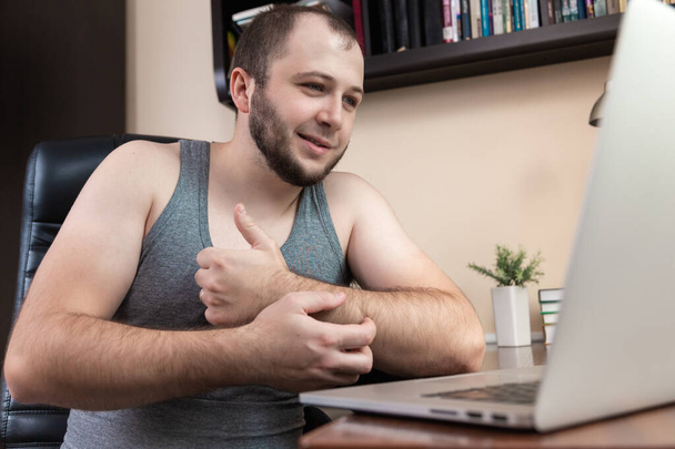 Досуг с помощью ноутбука. Молодой бородатый парень в домашней одежде серая футболка, использует ноутбук для просмотра видео, игры, общения, интернет-троллинг, покупки в Интернете во время коронавируса - Фото, изображение