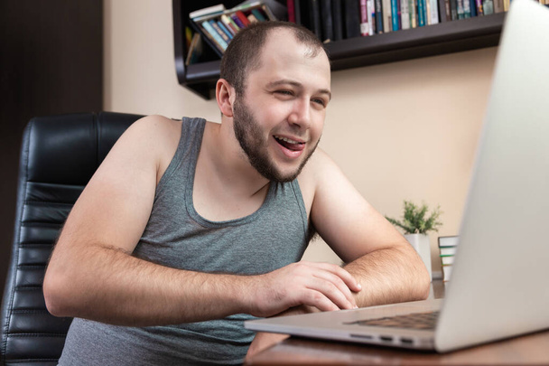 ノートパソコンを使ったレジャー。家庭用のグレーのTシャツを着た若いひげを生やした男は、ビデオを見るためにラップトップを使用し、ゲームをし、通信し、インターネットの回転、コロナウイルス中のインターネット上でのショッピング - 写真・画像