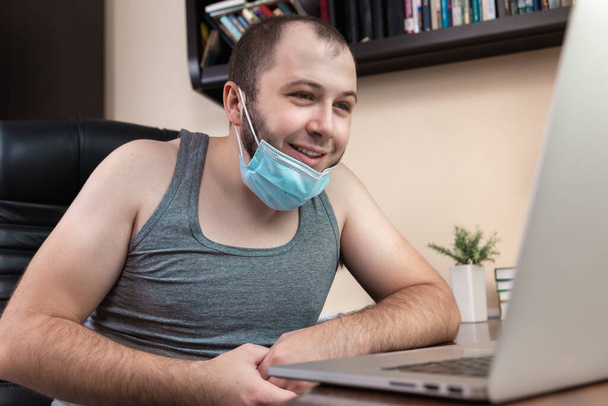 Досуг с помощью ноутбука. Молодой бородатый парень в маске для лица в домашней одежде использует ноутбук для просмотра видео, игр, общения, интернет-троллинга, покупок в Интернете во время коронавируса - Фото, изображение