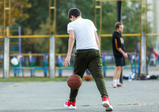 Cute young boy plays basketball on street playground. Teenager in white t shirt with orange basketball ball outside. Hobby, active lifestyle, sport activity for kids. - Photo, Image
