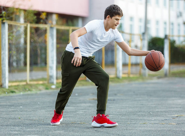 Lindo niño juega baloncesto en el patio de la calle. Adolescente en camiseta blanca con pelota de baloncesto naranja afuera. Aficion, estilo de vida activo, actividad deportiva para niños. - Foto, imagen
