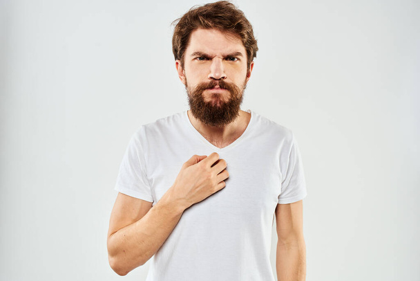Emotional bearded man in a white t-shirt gestures with his hands light background - Foto, Imagen
