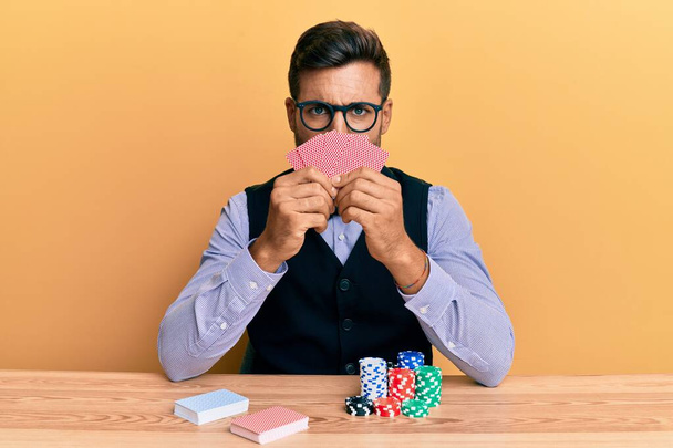 Красивый латиноамериканец крупье сидит на столе с фишками для покера и картами скептически и нервно, хмурясь расстроен из-за проблемы. отрицательный человек.  - Фото, изображение