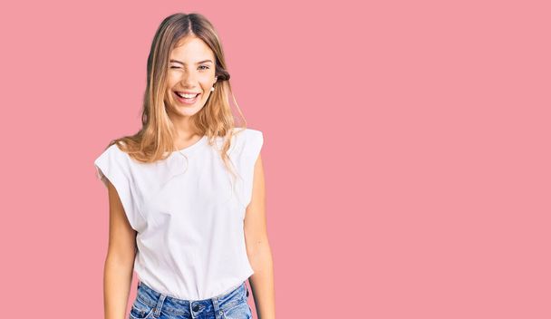 Beautiful caucasian woman with blonde hair wearing casual white tshirt winking looking at the camera with sexy expression, cheerful and happy face.  - 写真・画像
