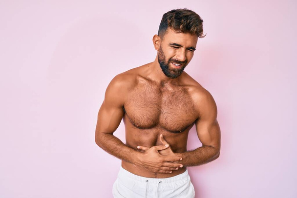 Young hispanic man standing shirtless with hand on stomach because nausea, painful disease feeling unwell. ache concept.  - Photo, Image