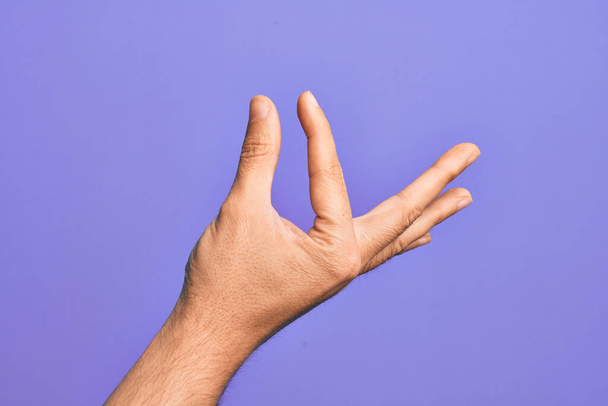 Mão de um jovem caucasiano mostrando dedos sobre fundo roxo isolado pegando e tomando coisa invisível, segurando objeto com os dedos mostrando espaço - Foto, Imagem