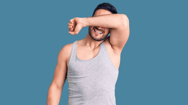 Young handsome man wearing swimwear and sleeveless t-shirt covering eyes with arm smiling cheerful and funny. blind concept.  - Photo, Image