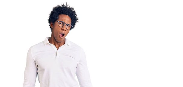 Knappe Afro-Amerikaanse man met afrohaar in casual kleding en bril in shock gezicht, op zoek sceptisch en sarcastisch, verrast met open mond  - Foto, afbeelding